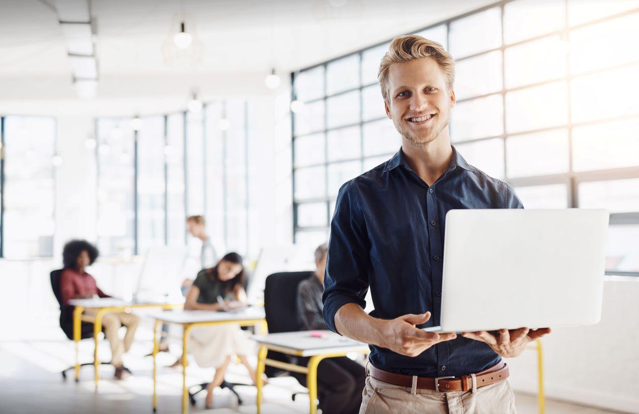 Lächelnder Mann mit Laptop in einem lichtdurchfluteten Büro, im Hintergrund ein Team bei der Arbeit – symbolisch für Zusammenarbeit und die Vorteile eines ERP-Systems wie Sage.