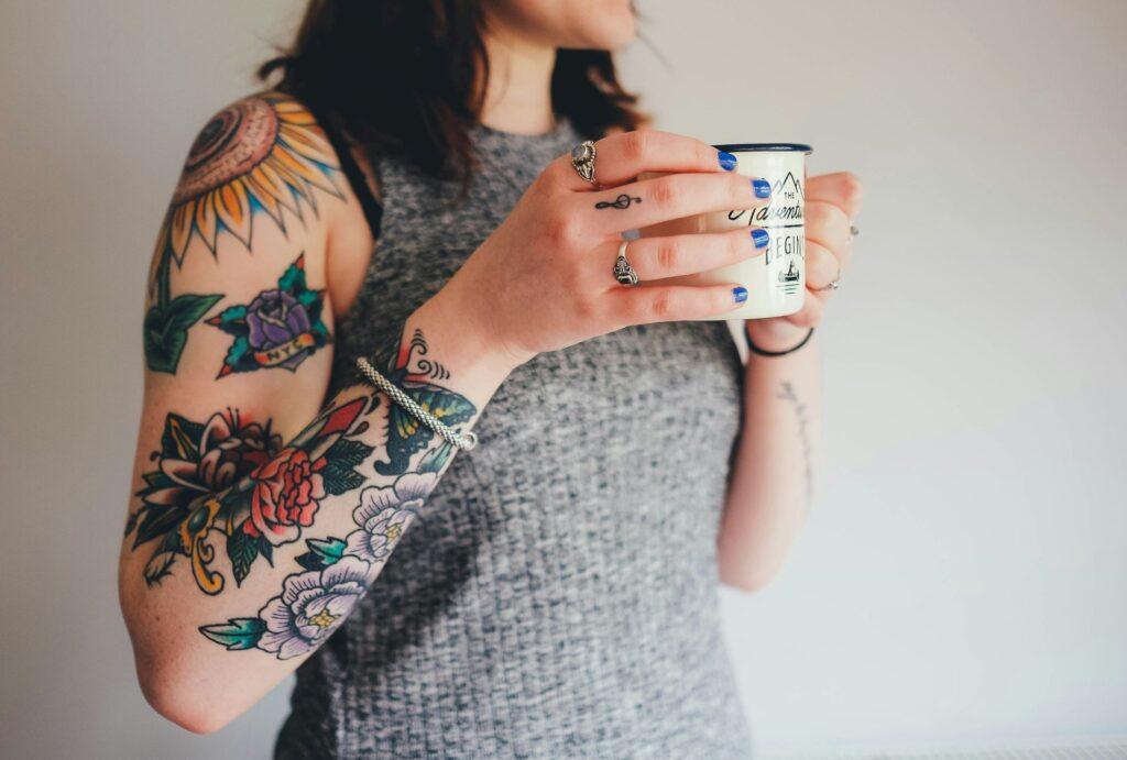 Dieses Bild zeigt eine Person mit bunten Tattoos auf dem Arm, die eine Tasse hält. Die Tattoos umfassen verschiedene Motive und Farben, wie Blumen und geometrische Designs.