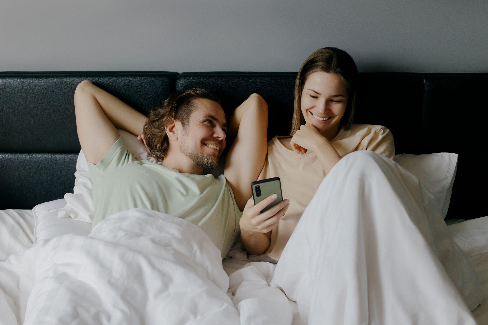 Paar entspannt auf Boxspringbett und schaut gemeinsam aufs Smartphone