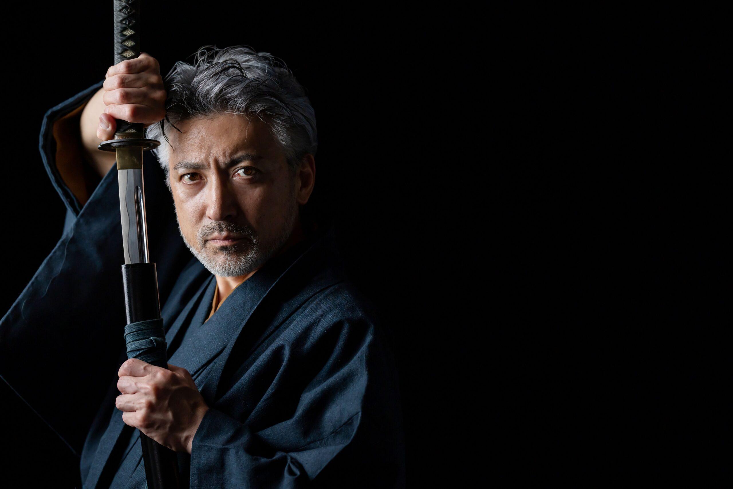 Katana in den Händen eines erfahren wirkenden Mannes vor dunklem Hintergrund
